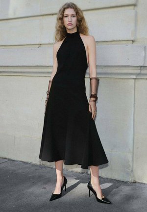 Vestido negro sin mangas en forma de halter con falda acampanada, hecho de una tela suave; combinado con tacones negros puntiagudos y acentos de brazo marrón traslúcido.