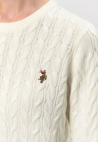 Close-up of a white cable-knit sweater with embroidered figure of a polo player on horseback in red and brown on the chest.
