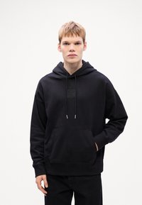 Zwarte katoenen hoodie met een voorzak, trekkoordkap en een gestructureerd embleem op de borst. Regular fit met geribde manchetten.