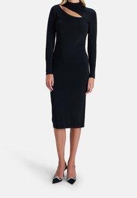 Abito lungo in maglia nera con scollo sulla spalla, design aderente e lunghezza al ginocchio. Abbinato a scarpe col tacco nere.