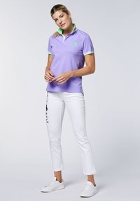 Lila Polohemd mit grünen Kragenakzenten und Logo, kombiniert mit weißen Hosen, die ein schwarzes Grafikdesign aufweisen. Das Modell steht vor einem neutralen Hintergrund.