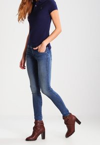 Námořnické polo tričko, slim-fit modré džíny, hnědé kotníkové boty s blokovým podpatkem a dekoračním páskem. Hladké textury a přiléhavý design.