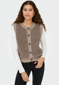 Brauner gestrickter Weste mit einer fusseligen Textur, mit Knopfverschluss, gerippten Kanten und grünen Knöpfen, getragen über einem weißen Langarmoberteil.