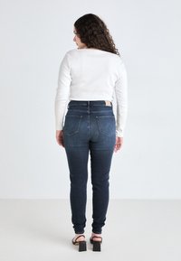 Σκούρες μπλε skinny τζιν παντελόνες με ψηλή μέση, πίσω τσέπες και ελαφριά υφή, συνδυασμένες με ένα λευκό μπλουζάκι μακρυμάνικο και ψηλοτάκουνα σανδάλια.