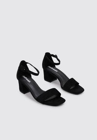 Sandalias de tacón de ante negro con tira en el tobillo, punta abierta y tacón cuadrado, con un forro interior suave y detalles de diseño mínimos.