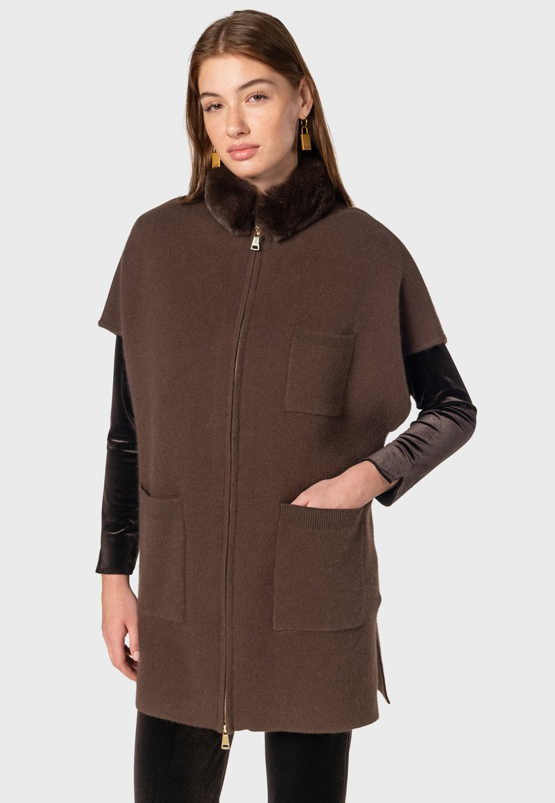 Gilet en laine marron avec col en fourrure, fermeture éclaire et deux poches avant ; présente une silhouette arrondie et des manches courtes.