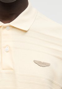 Μπλούζα πόλο σε κρεμ χρώμα, με υφή υφάσματος με οριζόντιες ρίγες, γιακά με τρία κουμπιά και μπεζ λογότυπο Aston Martin.