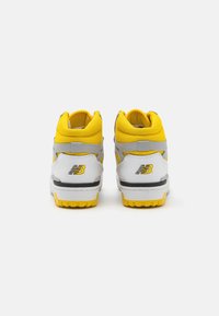 Scarpe sportive bianche e gialle con colletto imbottito, dettagli grigi e suole testurizzate. Presentano un marchio audace sul tallone.
