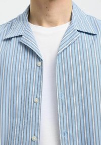 Světle modrá košile s vertikálními bílými a námořnickými pruhy, s špičatým límcem a dvěma knoflíky, vrstvená přes bílý tričko.
