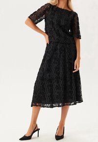 Sort blonde to-delt outfit med en kortærmet top med blomstermønster og en midi-nederdel. Glat tekstur med bølgede kanter.