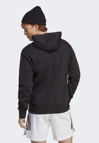 Black hooded sweatshirt with a soft texture, featuring a kangaroo pocket and ribbed cuffs. Worn with white shorts and a black beanie.