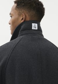 Mörkgrå ullrock med hög krage och en etikett som visar "M 73" på baksidan. Har sömlösa axlar och en strukturerad yta.