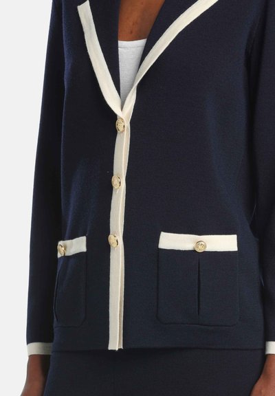 Cardigan bleumarin cu garnitură crem, nasturi aurii și două buzunare frontale. Realizat dintr-un material tricotat texturat. Design clasic cu guler.