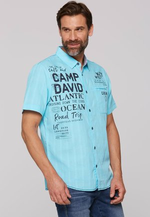 Helles blaues kurzärmliges Hemd mit Knopfleiste, das Textdrucke wie "CAMP DAVID" und "Road Trip" aufweist, mit einem strukturierten Stoff und schwarzen Akzenten.