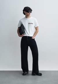 Λευκό T-shirt με το κείμενο "KIDSUPER", μαύρο τζιν με φαρδύ πόδι, μαύρη τσάντα και μαύρα παπούτσια με χοντρή σόλα. Διαθέτει μοντέρνα γυαλιά ηλίου και υφασμάτινο υλικό με υφή.