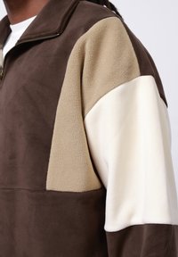 Fleece bunda s krátkym zipsom v hnedej, béžovej a krémovej farbe. Obsahuje dizajn s blokmi farieb, mäkkú textúru a viditeľný zips na golieri.