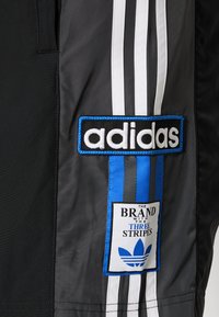 Black athletic track pants featuring three white stripes down the sides. Includes blue and black branded patches with text and a logo.
