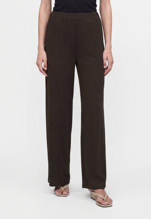 Personne portant un pantalon large marron foncé et des sandales à talons beige clair, debout devant un fond uni.