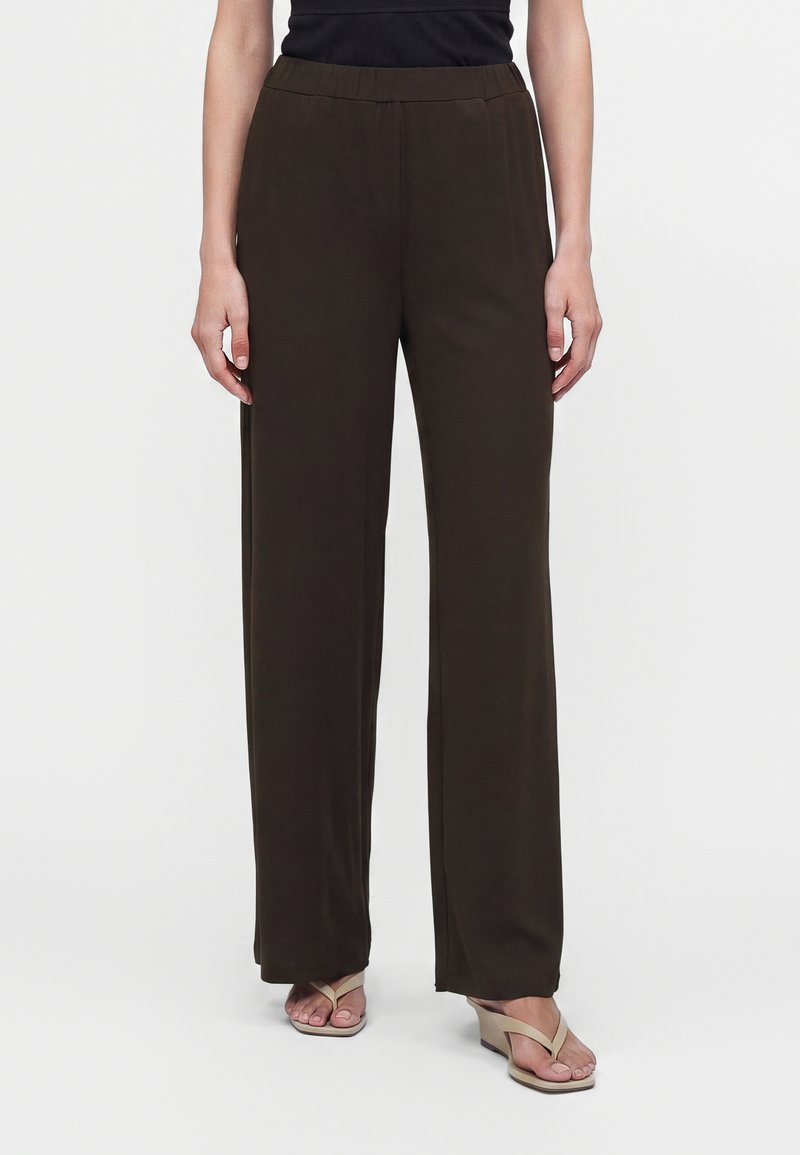 Personne portant un pantalon large marron foncé et des sandales à talons beige clair, debout devant un fond uni.