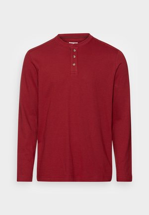 Maglietta Henley a maniche lunghe rossa con tre bottoni marroni e una sottile texture a righe orizzontali su uno sfondo chiaro.