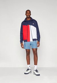 Chaqueta con cremallera en azul marino, rojo y blanco con diseño de bloques de color, combinada con pantalones cortos azul claro. Tejido suave y corte deportivo.