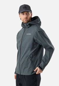 Muškarac u sivoj vodootpornoj jakni s kapuljačom i džepovima na patentni zatvarač, te s crnom bejzbol kapom, gleda u stranu.
