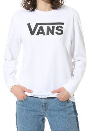 Osoba ubrana w białą koszulę z długim rękawem z czarnym logo "VANS" na piersi i niebieskie dżinsy, ręce w kieszeniach.