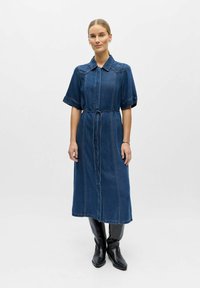 Femme debout portant une robe en jean mi-longue à manches courtes, une ceinture nouée à la taille, une broderie florale sur le col, et des bottes noires montantes jusqu'aux genoux.