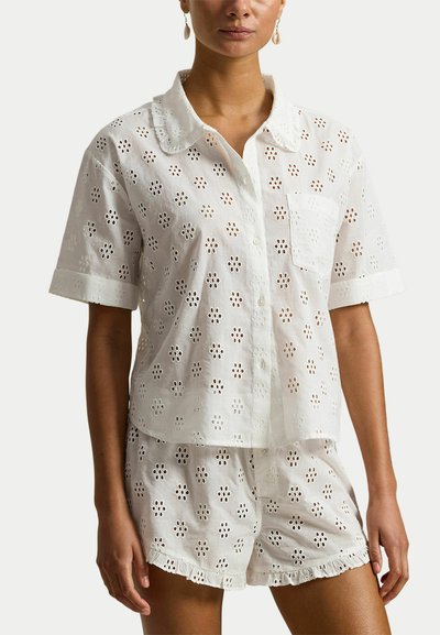 Vrouw draagt een witte blouse van oogjesstof met korte mouwen en een bijpassende korte broek met gerafelde randen.