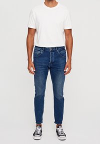 Blue denim jeans with a slim fit, five pockets, and subtle fading; paired with a plain white crew neck t-shirt and black sneakers.