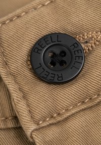 Brown fabric with a textured weave, featuring a black plastic button engraved with "REELL" and four holes for sewing.