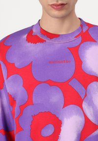 Marimekko hettegenser med store røde og lilla blomster mønstre, rund hals og myk tekstur. Merkedetaljer i rødt.