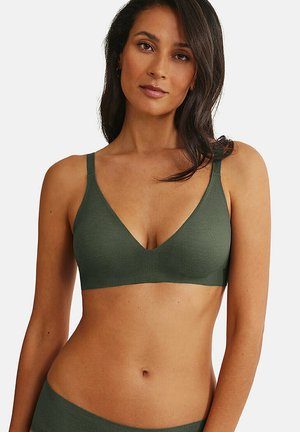 Femme avec un teint de peau moyen et de longs cheveux foncés portant un soutien-gorge vert olive sans coutures et une culotte assortie sur fond blanc.