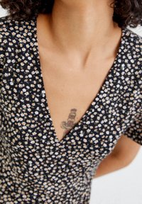 Schwarzes Kleid mit kleinem weißen und gelben Blumenmuster, V-Ausschnitt, kurzen Ärmeln und weichem Stoff. Auffälliges Tattoo auf der Brust sichtbar.