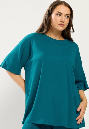 Frau trägt ein locker sitzendes, türkises Kurzarmshirt mit rundem Ausschnitt und posiert mit einer Hand auf der Hüfte vor einem schlichten Hintergrund.