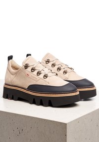 Scarpe in suede beige con punta in gomma nera e suola spessa. Presentano occhielli metallici e lacci piatti. Dettaglio del logo visibile sul lato.