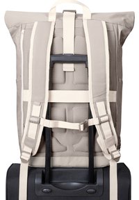 Backpack with adjustable beige fabric straps, padded mesh backing, roll-top design, and a grip handle, mounted on a black wheeled suitcase.
