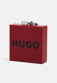 Boutons de manchette en argent avec le logo gravé "HUGO" sur une boîte rouge comportant un texte noir en lettres majuscules. La boîte est rectangulaire avec une finition lisse.