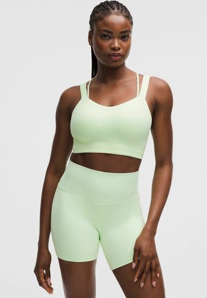 Mujer con trenzas, vistiendo un sujetador deportivo ajustado verde claro y pantalones cortos de ciclista de cintura alta, de pie frente a un fondo liso.