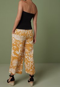Donna che indossa pantaloni a gamba larga giallo senape con stampa floreale bianca, top senza spalline nero e sandali con zeppa neri, in piedi davanti a uno sfondo verde.