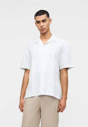 Noor mees, kes kannab lühikeste varrukatega valget triibulist nööbitavat särk ning beeže pükse, seisab ühevärvilise heleda tausta ees.