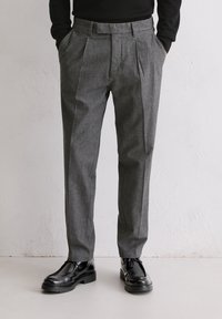 Graue, gefaltete Hose aus strukturiertem Stoff, mit einem maßgeschneiderten Schnitt und seitlichen Taschen. Kombiniert mit schwarzen Lederschuhen.