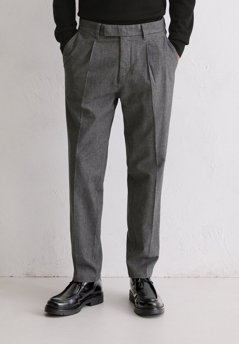 Grijze geplooide broek van textuurstof, met een getailleerde pasvorm en zijzakken. Gecombineerd met zwarte leren schoenen.
