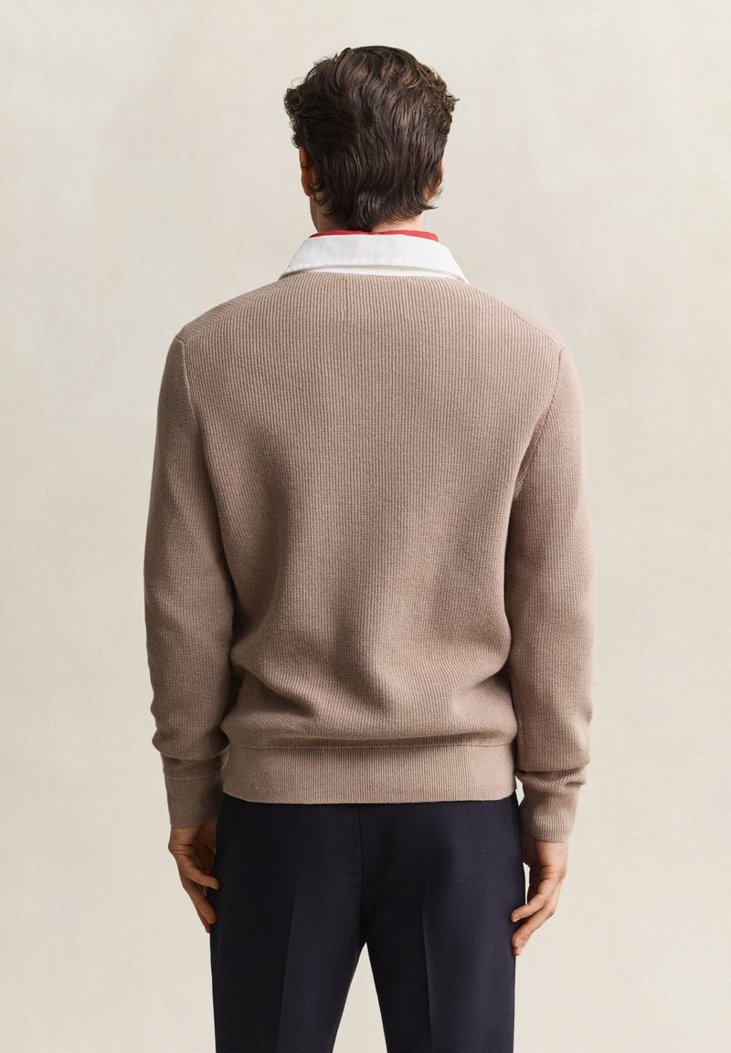 Man facing away wearing a beige ribbed sweater over a white collared shirt with navy trousers against a plain background.