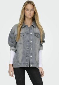 Giacca di jeans grigia oversize con chiusura a bottoni, due tasche sul petto e maniche arrotolate. Abbinata a una camicia bianca aderente sotto.