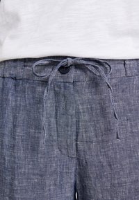 Lněné kalhoty v tmavě modré barvě s texturovanou vazbou. Mají pásek se skořicovým stahováním a jediné knoflíkové zapínání vpředu.