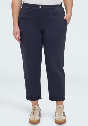 Henkilö pukeutuneena laivastonsinisiin, käännettyihin housuihin ja beigen värisiin Adidas-tossuihin, joissa on mustat raidat, seisoo yhdellä kädellä taskussa.