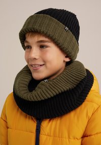 Stickad mössa och matchande halsduk i olivgrön och svart. Halsduken har en bred ribbad textur. Klart gul puffjacka i bakgrunden.