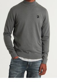 Gray knit sweater with a high ribbed collar, featuring a small black logo patch. The fabric has a smooth texture, paired with blue jeans.