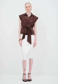 Blusa marrone a maniche corte con dettaglio a nodo davanti, abbinata a una gonna a tubino bianca e sandali neri con tacco. Tessuto liscio e setoso.